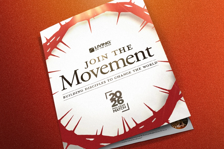 A mock-up of a printed brochure lying on a textured orange surface. The white cover features the "LIVING ON THE EDGE" logo and text that reads "JOIN THE Movement: BUILDING DISCIPLES TO CHANGE THE WORLD," encircled by a red crown of thorns design with the "2026 MID-YEAR MATCH" logo at the bottom.