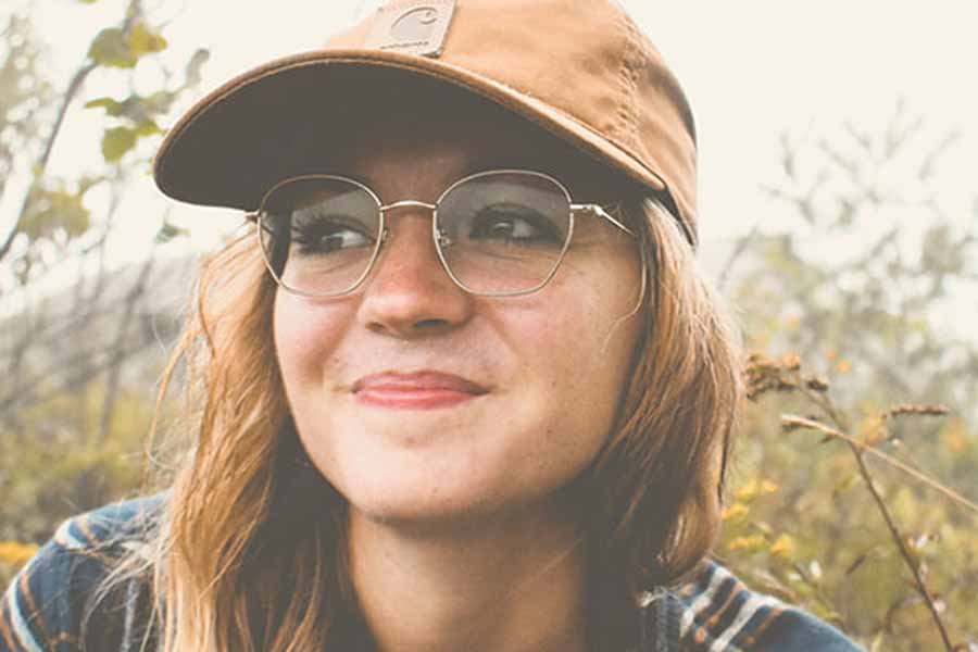 A smiling young woman wearing a brown Carhartt baseball cap, wire-rimmed glasses, and a plaid shirt outdoors.