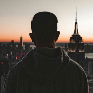 Silhouette of a person wearing a hoodie, looking out over the New York City skyline and the Empire State Building at sunset.