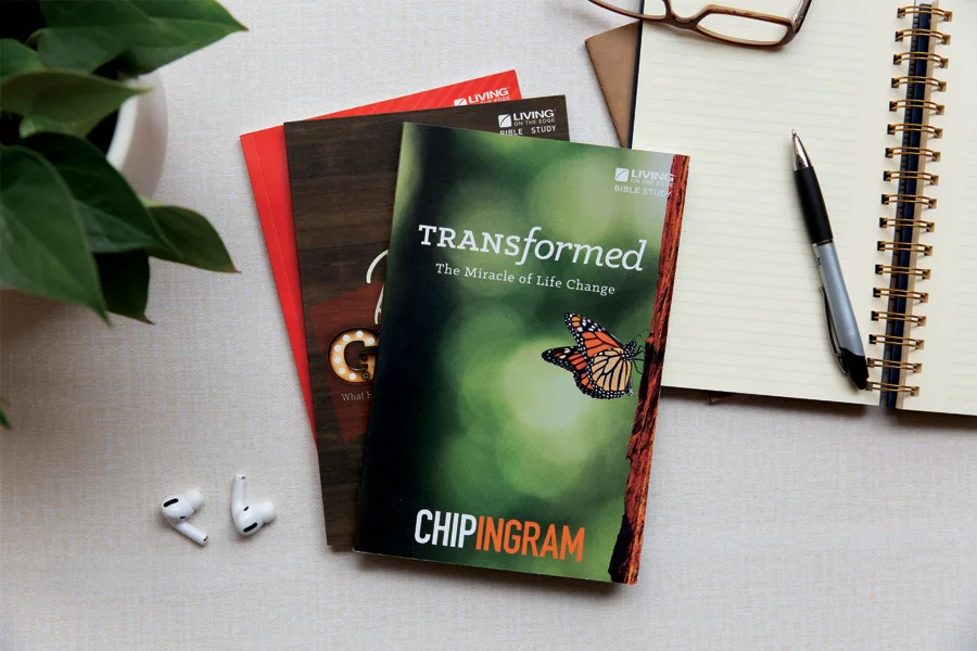 Three "Living on the Edge" Bible study guides, including "Transformed" by Chip Ingram, on a desk with a notebook, pen, and earbuds.