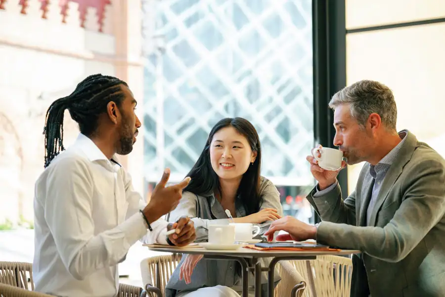 Three friends having a discussion at a cafe table. A man gestures while speaking, a woman listens with a smile, and another man drinks from a coffee cup.