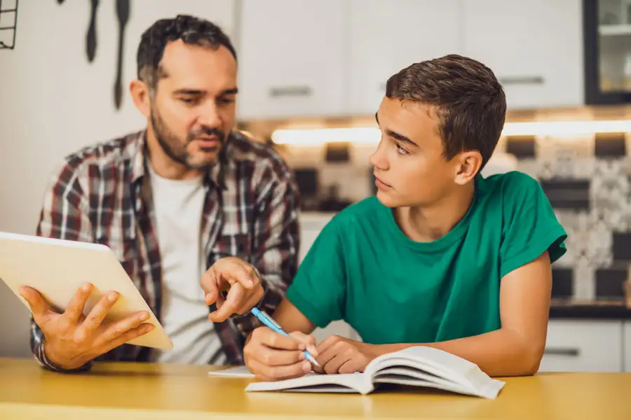 A teen boy and his dad studying at a table with an open textbook