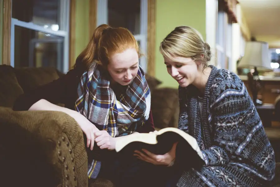 Two young women reading a book together on the couch.