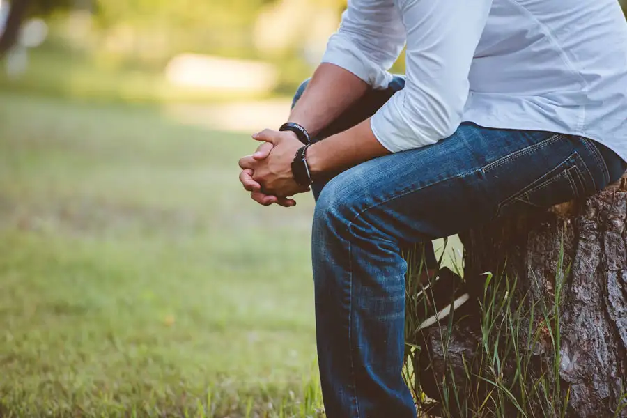 Close-up of a man sitting on a tree stump outdoors with his hands clasped together in prayer.