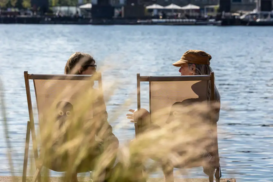 Two people relaxing on outdoor lounge chairs facing each other near the water's edge, talking on a sunny day.