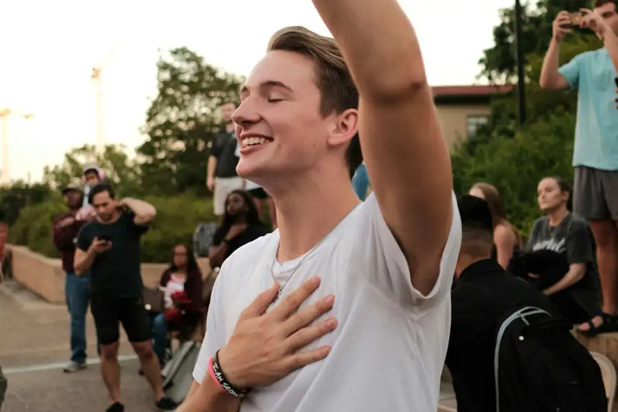 A smiling young man in a white T-shirt standing outdoors with his eyes closed, placing one hand over his chest and raising the other hand in worship.