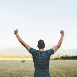A man seen from behind raising both arms in victory while standing in an open field facing mountains.