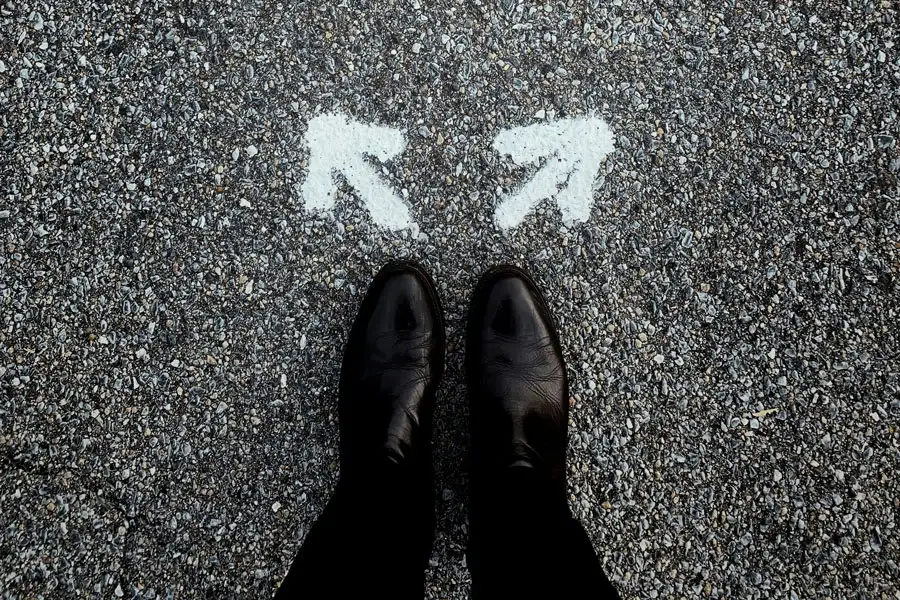 Top-down view of someone wearing black leather shoes standing on asphalt in front of two white painted arrows pointing left and right.