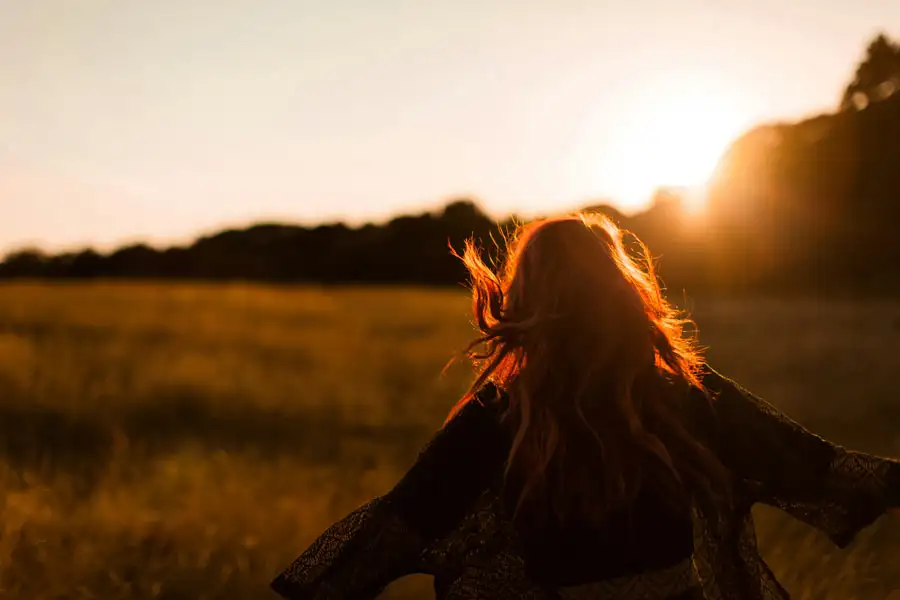 view of a woman with red hair flowing in the wind, walking through a field toward a warm sunset.