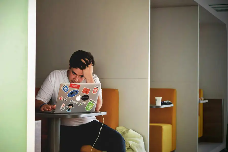 A young man sitting in an office booth, resting his head in his hand with a concentrated, stressed expression as he types on a laptop.
