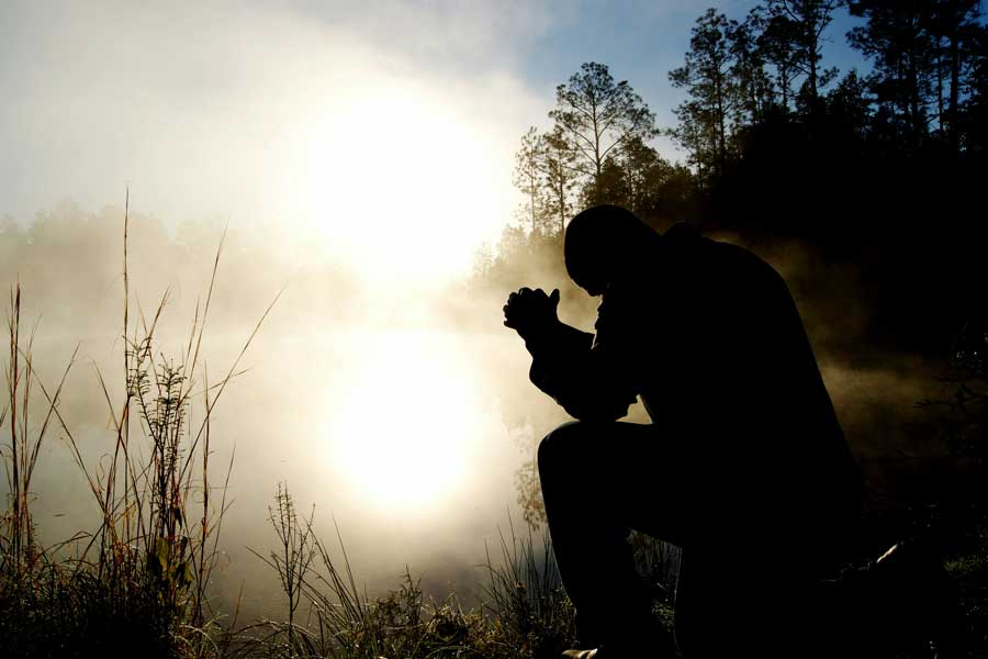 Silhouette of a person kneeling in prayer with folded hands beside a misty lake at sunrise.