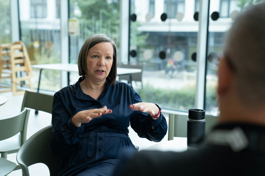 A woman in a navy blue dress gesturing with her hands while speaking to a person sitting across from her indoors.