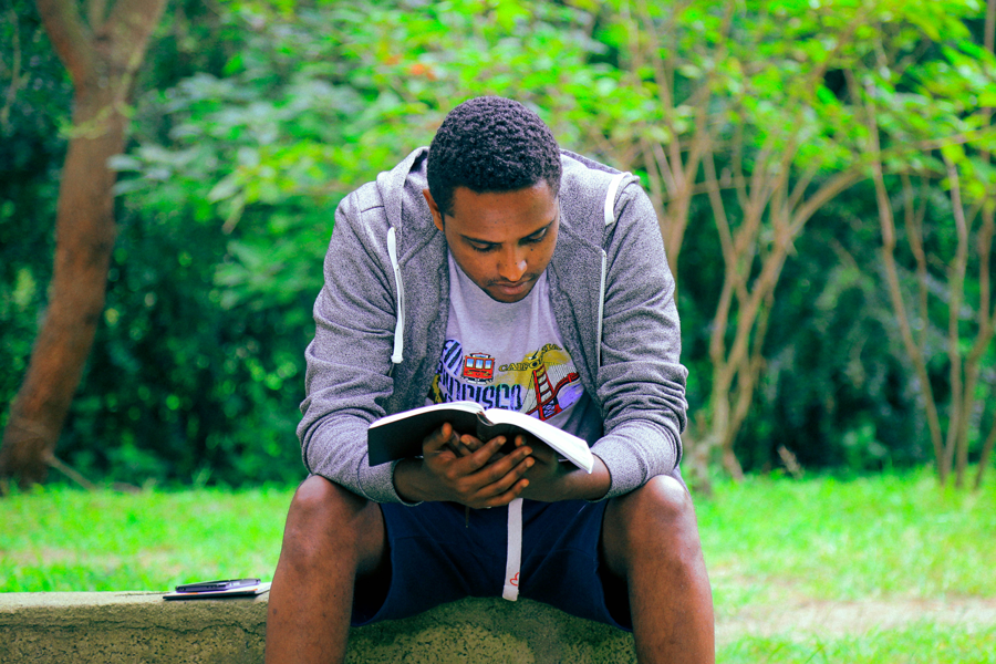 : A young man wearing a grey hoodie sitting outdoors, reading an open book while leaning forward against a lush green background.