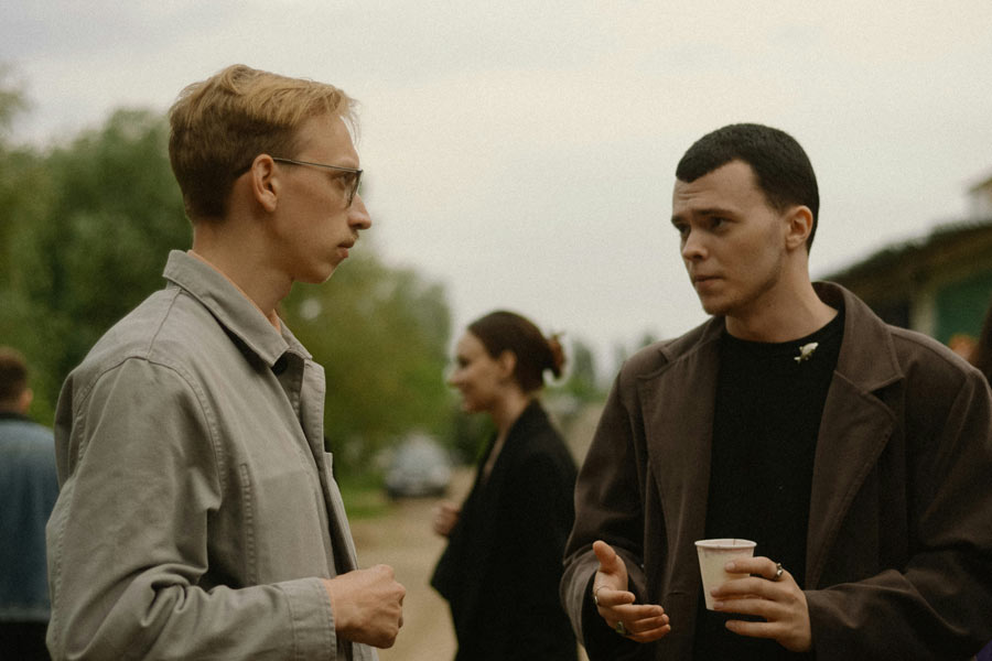 Two men having a serious conversation outdoors, with one holding a paper cup and gesturing as he speaks.