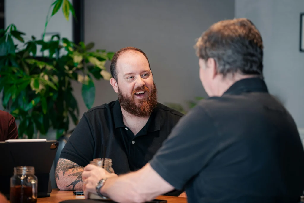 A bearded man with arm tattoos smiling and laughing during a conversation with another man across a table indoors.