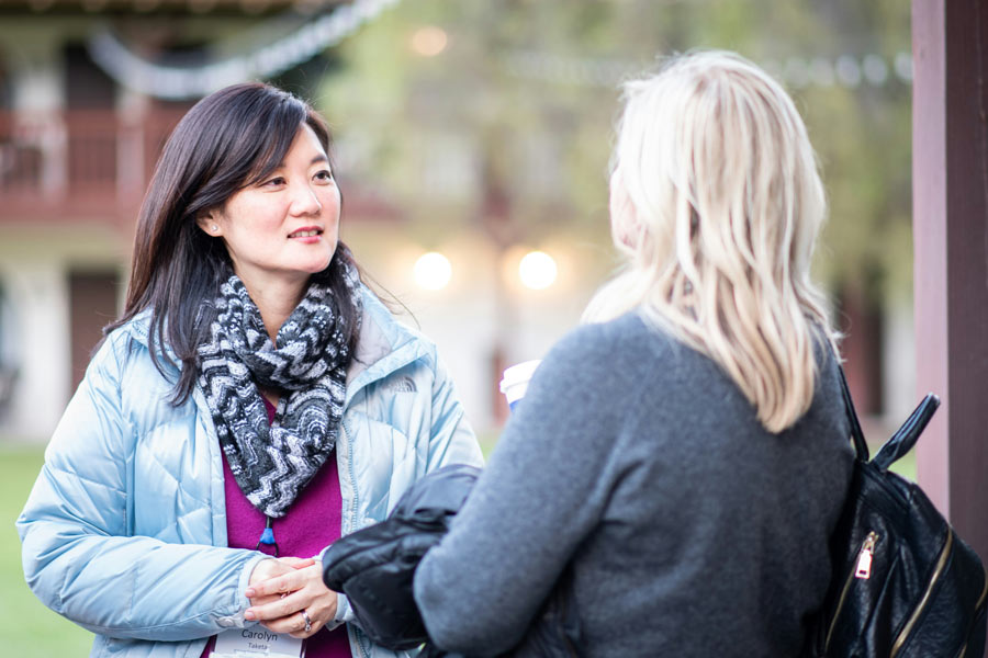 An Asian woman wearing a light blue jacket and patterned scarf listening intently to a blonde woman outdoors.