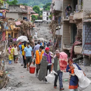 People walk through a debris-filled street carrying supplies and belongings past heavily damaged multi-story buildings after a flood or natural disaster.