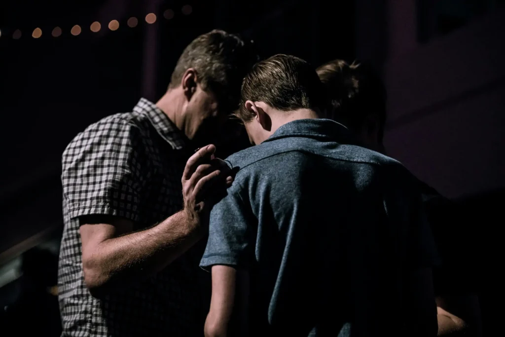 A man placing his hand on another person's shoulder as they bow their heads together in prayer.