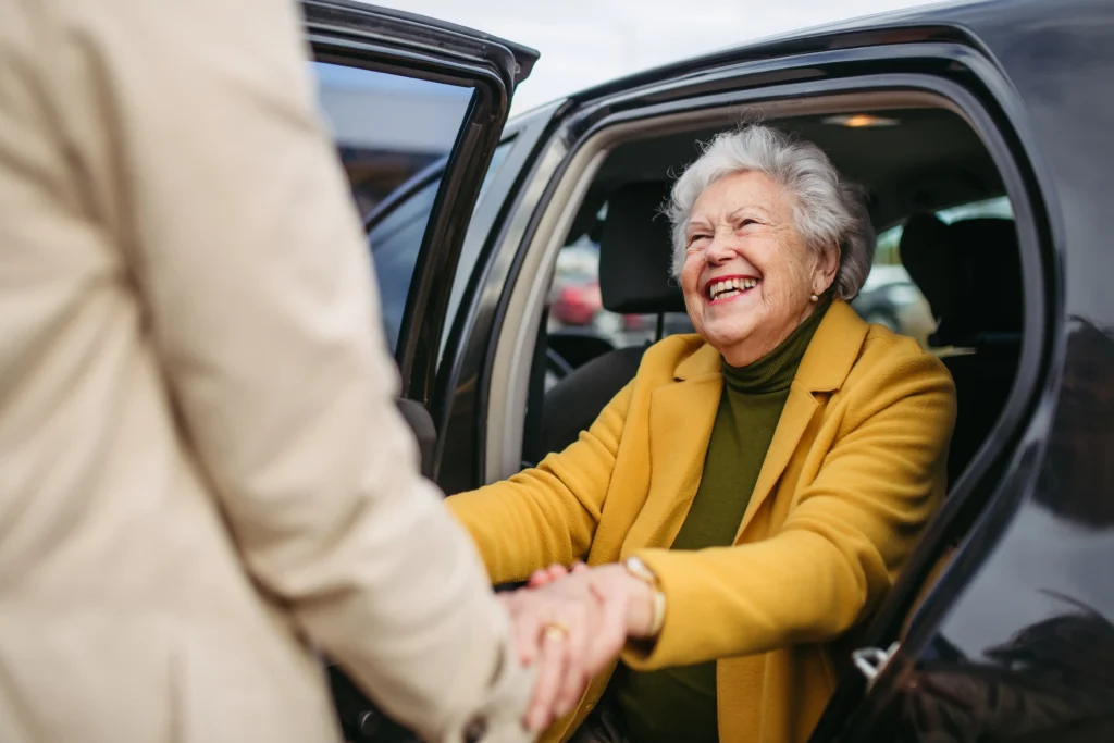 A smiling elderly woman in a yellow jacket sitting in the open door of a car, holding hands with someone standing outside to help her. 
