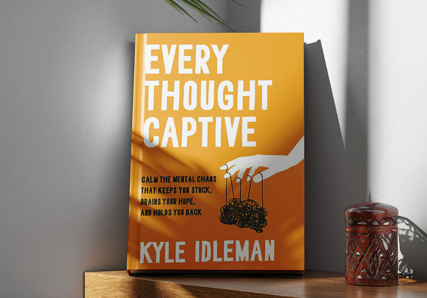 Orange book titled "Every Thought Captive" by Kyle Idleman on a wooden shelf. The cover shows a white hand holding strings attached to a tangled scribble.